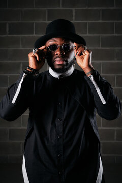 Stylish African American male in black apparel putting on trendy round sunglasses and looking at cmaera while standing on background of brick wall in studio