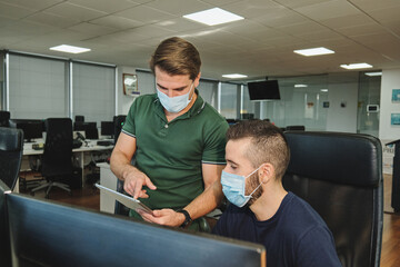 Serious male colleagues in medical masks discussing project while working on software development in modern office