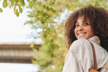Low angle side view of charming curly haired ethnic female in coat and with backpack standing in garden in fall and looking away