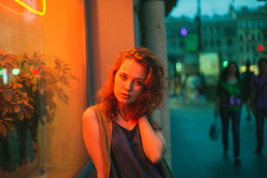 Attractive sensual young red haired female leaning on wall of urban building with neon illumination and looking at camera while standing on street of Saint Petersburg in evening time