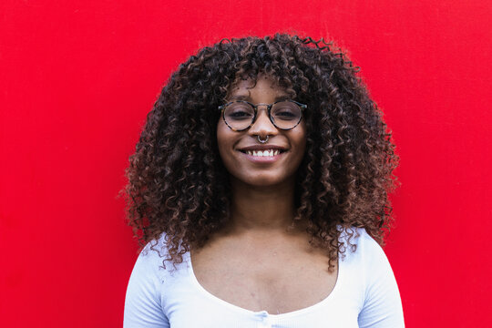 Cheerful black woman with piercing and glasses smiling and looking at camera against bright red wall
