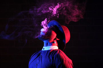 Side view of stylish African American male in hat and sunglasses standing on black background in studio with neon illumination and smoking vape while exhaling steam