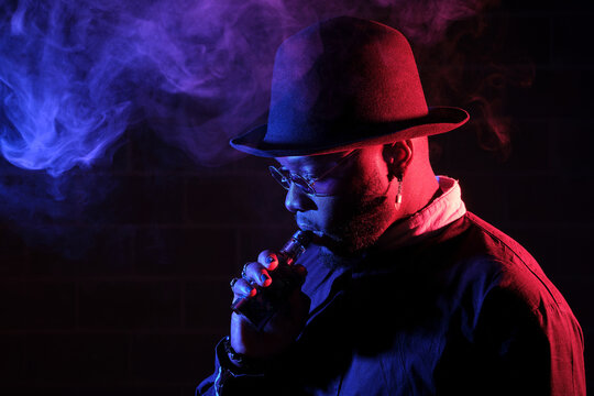 Side view of stylish African American male in hat and sunglasses standing on black background in studio with neon illumination and smoking vape while exhaling steam