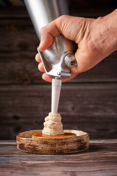 Unrecognizable chef using cream whipper and garnishing delicious cheesecake in plate on wooden table
