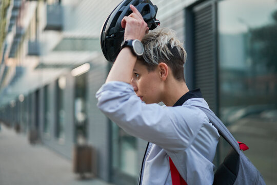 Young woman cyclist puts on helmet while standing next to modern building on summer day in city, side view