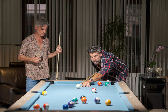 Happy mature friends in casual clothing playing pool and drinking cocktails while resting in cozy room in light of lamps
