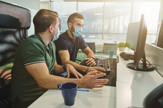 Male coworkers in medical masks sitting together using program for software during coronavirus pandemic