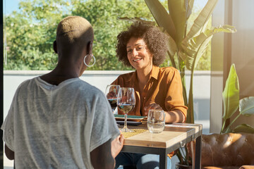 Friendly African American females sitting at table on terrace on cafe while drinking wine and laughing