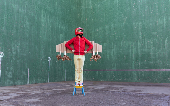 Unrecognizable playful kid wearing handmade carton rocket wings and helmet standing on chair while pretending being astronaut