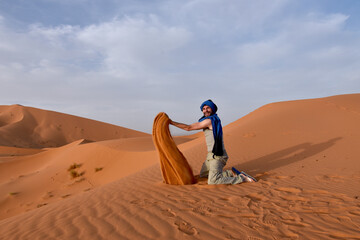 Kneeling occidental man throwing silky orange sand from the top of a sand dune in the desert, creating a sandy orange flame shape in the air