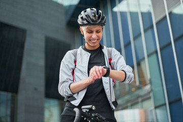 Young woman cyclist looks at wristwatch on background of modern building on summer day in city