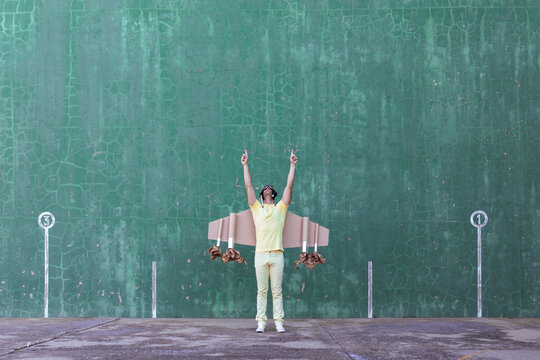 Cheerful male in handmade cardboard wings standing with raised arms and looking up while dreaming about achieving success