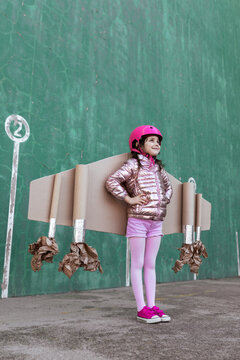 From below side view of cute girl in pilot helmet and handmade creative carton wings standing on street on green background looking away