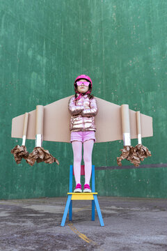Serious little girl in cardboard plane wings and helmet with funky sunglasses standing on chair on green background