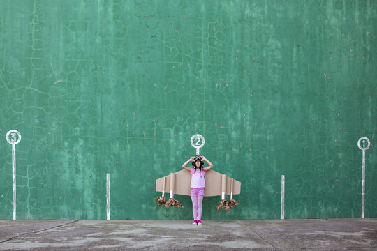 Cute girl in pilot helmet and handmade creative carton wings standing on street with raised arms looking up