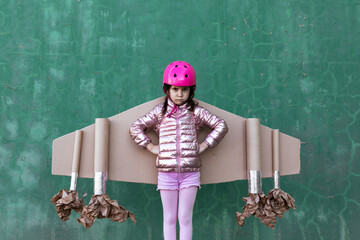 Discontent child in pink helmet and handmade cardboard airplane wings standing with hands on waist and looking at camera