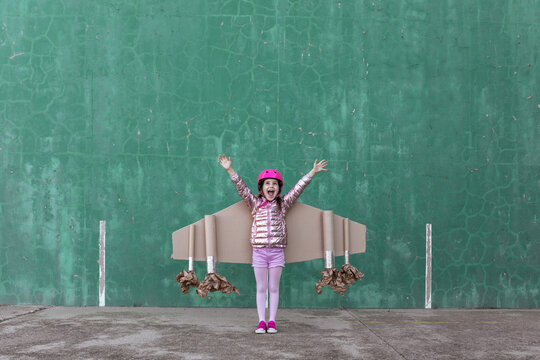 Cute girl in pilot helmet and handmade creative carton wings standing on street with raised arms looking up