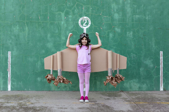 Cute girl in pilot helmet and handmade creative carton wings standing on street with raised arms and showing strength