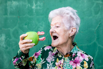 Stylish elderly female in trendy outfit holding green apple with denture while standing against green background