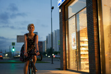 Woman in dress rides bicycle in night city near store