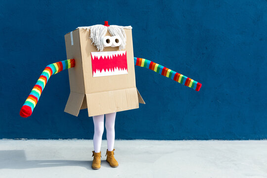 Unrecognizable playful kid wearing funny costume of monster made of carton box standing on street during holiday celebration