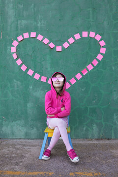 Cheerful cool little girl wearing stylish pink clothes and sunglasses sitting on chair near wall with heart made of sticky notes