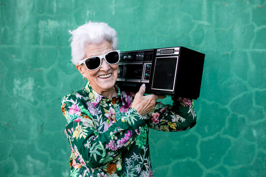 Happy elderly gray haired female in stylish outfit carrying record player on shoulder while standing against green wall on street looking at camera