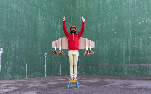 Unrecognizable playful kid wearing handmade carton rocket wings and helmet standing on chair with fists pointing up while pretending being astronaut