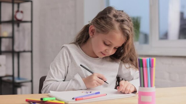 Cute little girl are drawing pictures with felt-tip pen on the white paper album sitting at the table. Playing alone, creative art activity at home. Spending free time at home during school holidays.
