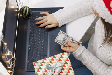 Woman in santa hat sits by laptop and holds dollar bill, concept of shopping or payment online at New Year or Christmas