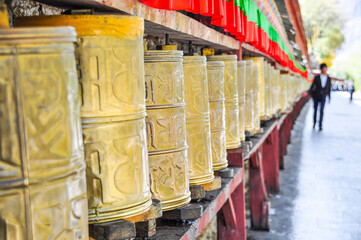 Fototapeta na wymiar Golden prayer wheels are lined up along a street in Lhasa, Tibet. The wheels have different sizes, heights and lettering. Focus on the wheels.
