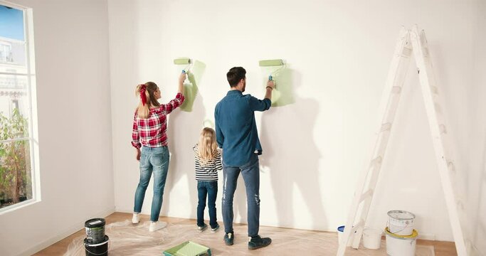 Rear of Caucasian cute family spending time together working in new apartment painting wall in green olive color using roller brush. Parent and little girl daughter renovating and redesigning kid room
