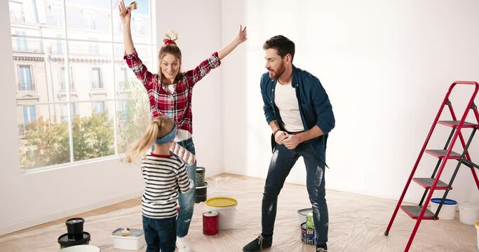 Cheerful lovely family having fun with little adorable girl child jumping, dancing and fooling around during renovation and home repair works. Parents and pretty daughter during room redesign
