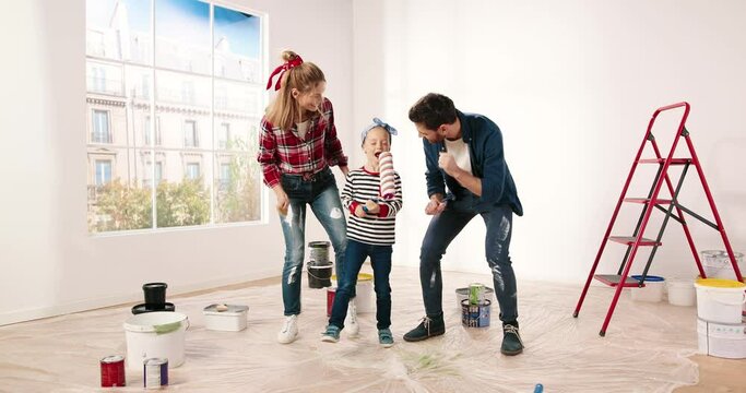 Happy smiling family having fun with little adorable girl dancing and fooling around during home repair in new apartment. Parents dance while small daughter singing into roller brush. Redesign concept