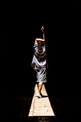 A woman in a skirt walks along a beam of light in the dark