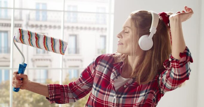 Close up of happy funny beautiful Caucasian female in headphones listening to music and singing song using roller brush as a microphone and musical instrument having fun during home repair