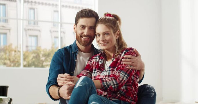 Close up of Caucasian cheerful young male and female couple sitting in room together, embracing, looking at camera and smiling in good mood. Family time. Happy wife and husband. Repair concept