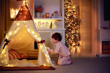 cheerful kid in pajamas playing with illuminated teepee tent at home on Christmas holidays © Olesia Bilkei