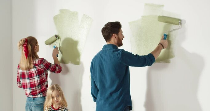 Back view of Caucasian happy nice family parents with little kid girl painting wall in green color using roller brush in hand in apartment renovating home. House redesigning and repair. House concept