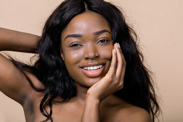 Attractive smiling African American lady with perfect skin and nude makeup, keeping one hand under...