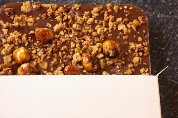 Milk chocolate bar with whole and cracked nuts in a white cardboard package with no brand name on a black background. Top view