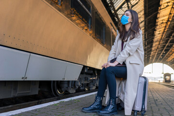 Fototapeta na wymiar Lonely sad girl in medical mask sits on suitcase waiting for train. Female traveler at empty train station