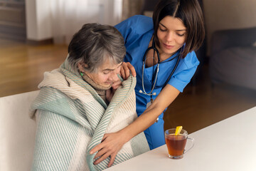 A young nurse is caring for an elderly 80 year old woman. The doctor brought hot lemon tea and sits with the patient, wrapped in a blanket when she has a cold. Trust and care, medicine