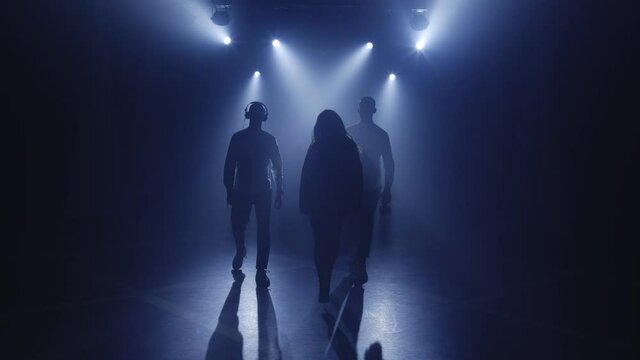 Backlit silhouettes of singer vocalist girl, saxophonist sax, dj man with headphones walking forward in dark nightclub disco for starting performance. Light appears and illuminates musical group band