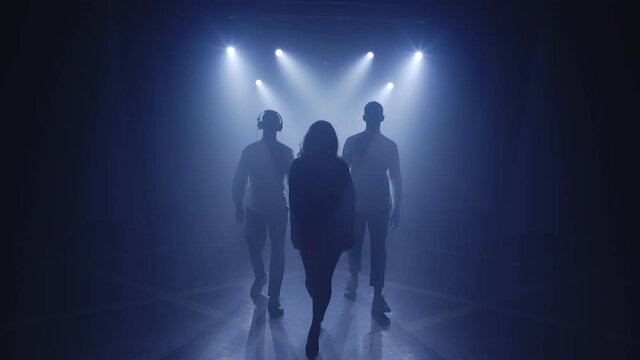 Backlit silhouettes of singer vocalist girl, saxophonist sax, dj man walking forward in dark musician nightclub disco for starting concert on stage. Light appears and illuminates musical group band