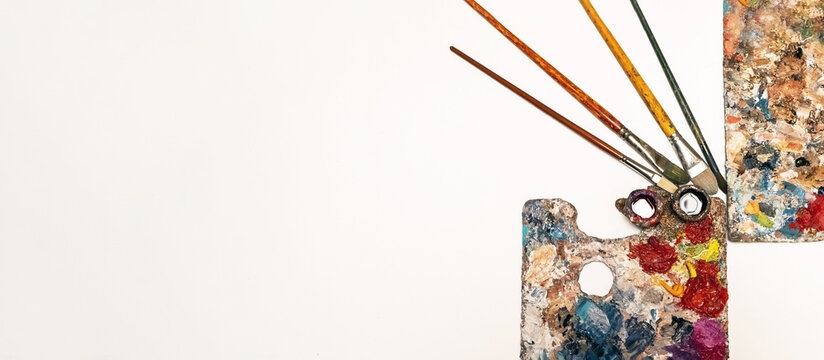 Art materials such as brushes of different sizes, palettes with vibrant embossed oil paints and a stretcher lie on a white background. View from above.