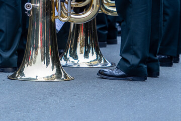 Brass band members stand on asphalt pavement during a parade. Their french horns and tubas are...