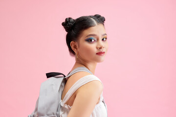 Fashionable schoolgirl girl holding a backpack behind her shoulders and looking directly at the camera isolated on shine pink background