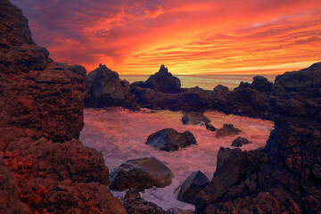 Ocean water fills up a rocky pool area on Pico Island in the Azores Islands during a beautiful sunset with a red, orange, yellow, and magenta sky.