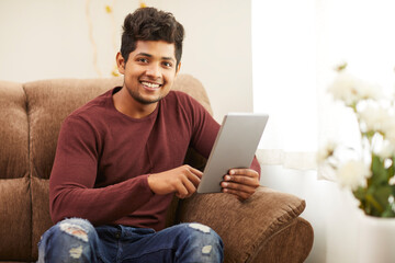 Handsome smiling young man using digital tablet on sofa at home.